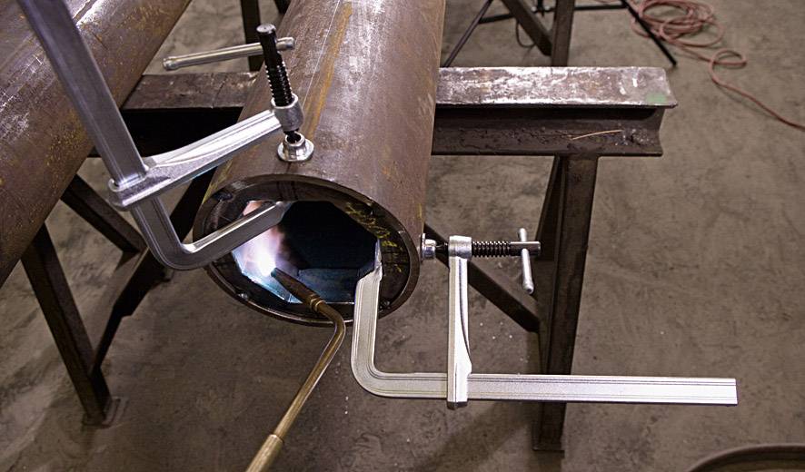 Eine Metallröhre wird mit Hilfe eines Brenners und Schraubzwingen auf einer Werkbank bearbeitet. Eine Flamme ist im Inneren sichtbar.