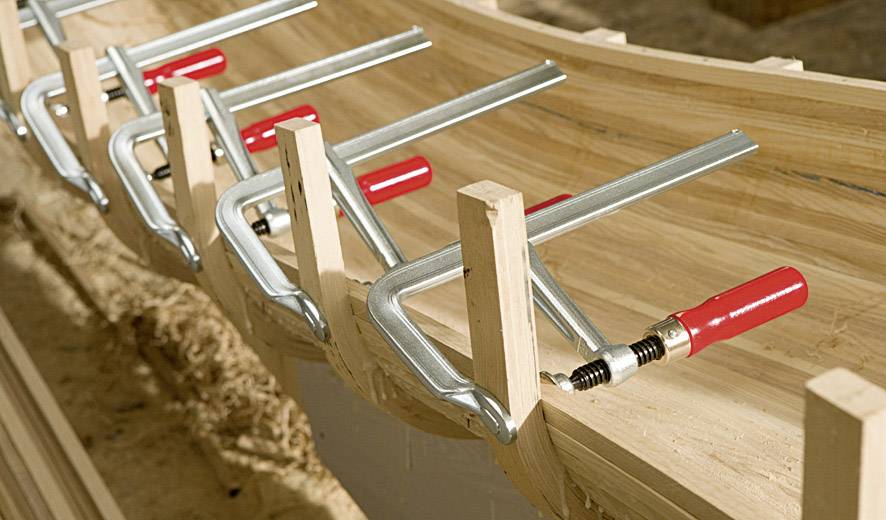 Mehrere Schraubzwingen halten gebogene Holzleisten zusammen. Sie dienen dazu, eine Struktur in einer Werkstatt zu formen oder zu fixieren.