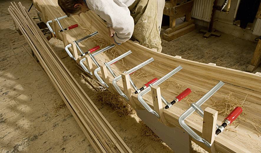 Eine Person bearbeitet ein Holzboot in einer Werkstatt, verwendet Klemmen, um die Planken zu befestigen. Holzspäne bedecken den Boden.