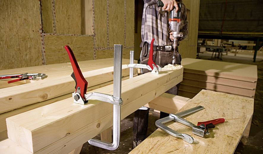 Ein Handwerker bearbeitet mit einer Bohrmaschine einen Holzbalken, der von zwei großen Zwingen fixiert wird. Im Hintergrund liegen Holzplatten.