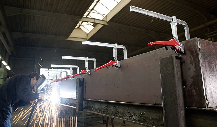 Ein Arbeiter schweißt in einer Werkhalle ein großes Metallstück, das mit mehreren roten Zwingen fixiert ist. Funken fliegen.