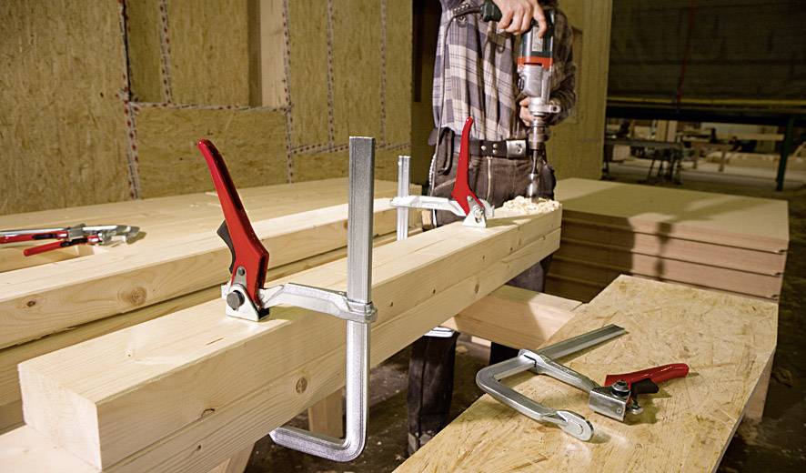 Arbeiter bearbeitet Holzbalken in einer Werkstatt mit Bohrer und Schraubzwingen, um die Holzkonstruktion zu fixieren.