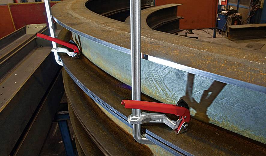 Zwei große Stahlplatten werden mit roten Schraubzwingen an einer Werkbank befestigt, um gebogene Stahlträger zu sichern.
