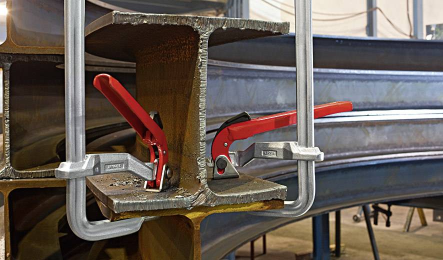 Zwei Metallträger werden von großen Schraubzwingen mit roten Griffen zusammengehalten. Die Umgebung deutet auf eine Werkstatt hin.