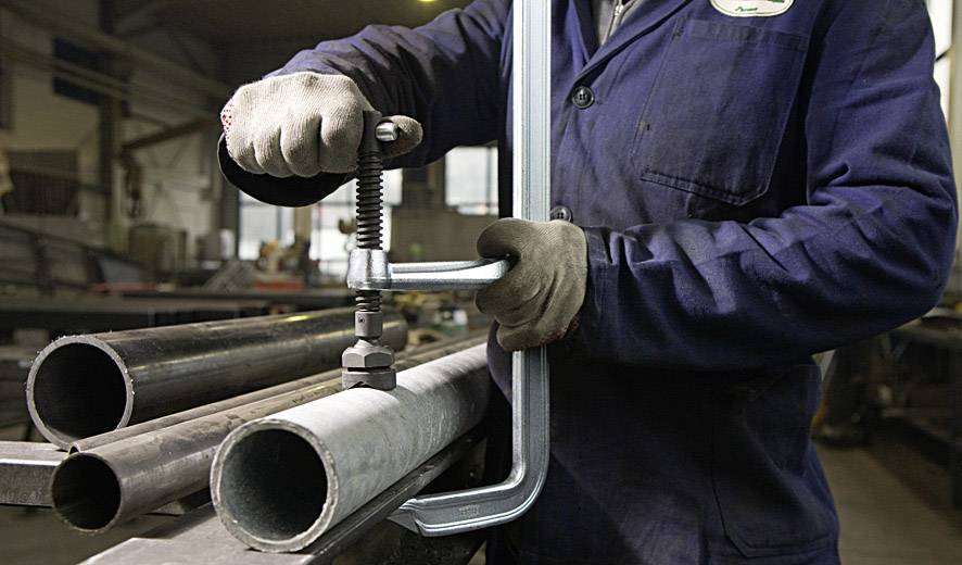 Eine Person in Arbeitskleidung verwendet eine C-Klemme, um Metallrohre in einer Werkstatt zu fixieren. Im Hintergrund sind weitere industrielle Werkzeuge sichtbar.