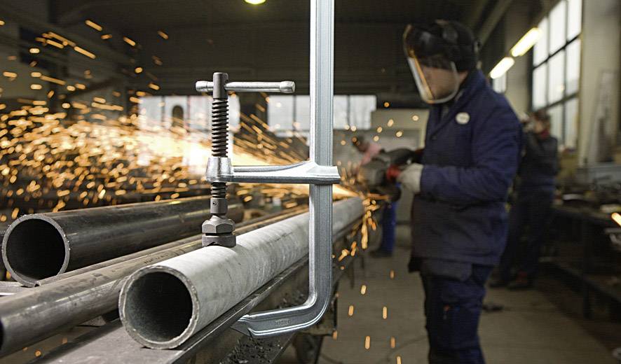 Eine Person schleift Metallrohre, während Funken fliegen. Im Vordergrund sind eine Klemme und Metallrohre zu sehen.