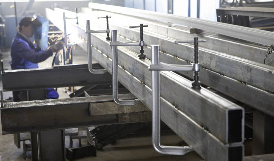 Eine Person schweißt in einer Werkstatt, Metallwerkstücke sind mit Klemmen befestigt und aufgereiht; Arbeitsprozess in der Metallverarbeitung.