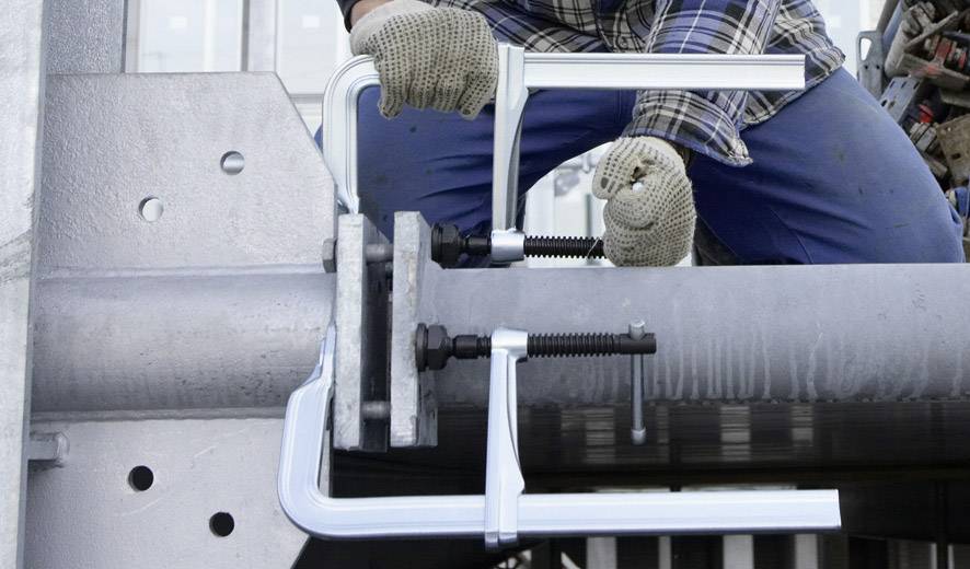 Eine Person in Arbeitskleidung zieht Bolzen an einem Metallkonstrukt fest, um zwei Bauteile mit Schraubzwingen zu verbinden.