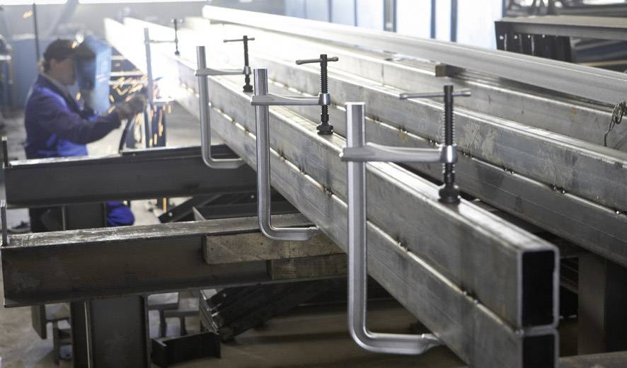 Eine Person schweißt Metall in einer Werkstatt; lange Metallbalken werden mit Schraubzwingen fixiert.