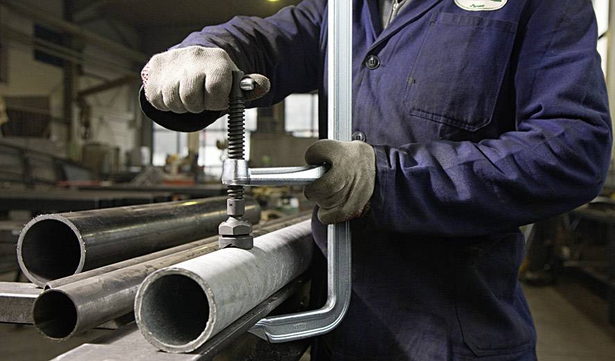 Eine Person spannt ein Metallrohr in einer Werkstatt mit einer Zwinge ein. Sie trägt Arbeitskleidung und Handschuhe.