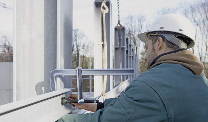 Ein Bauarbeiter mit Schutzhelm befestigt Metallbalken mit einer Klemme. Im Hintergrund sind Baugerüste und Bäume sichtbar.