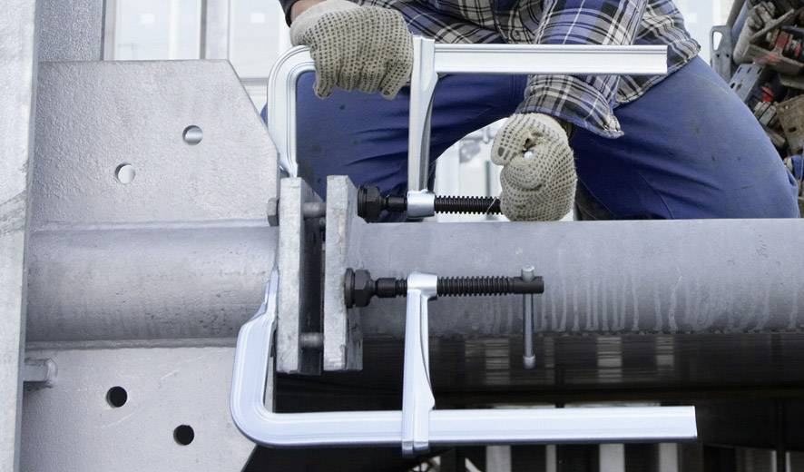 Eine Person befestigt mit Schraubzwingen ein Metallrohr an einem Gerüst. Sie trägt Arbeitskleidung und Handschuhe.