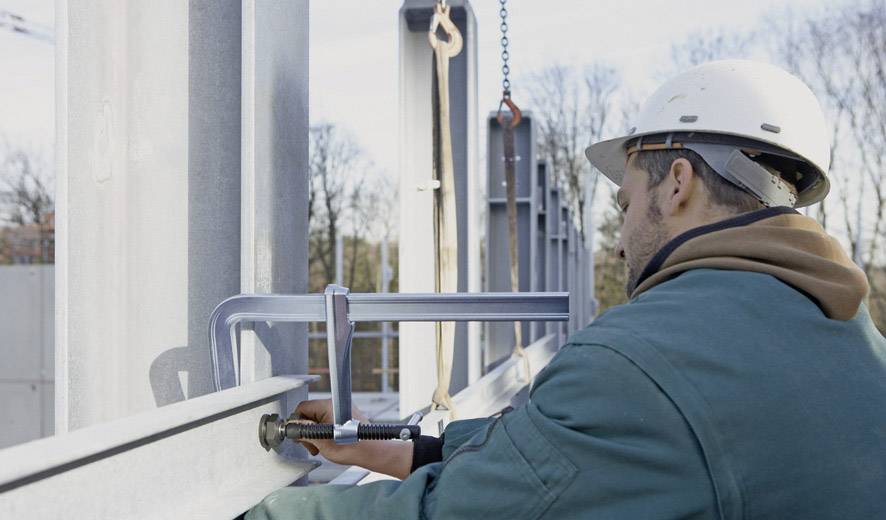 Ein Bauarbeiter mit Schutzhelm befestigt ein Metallträgerteil auf einer Baustelle mit einem Schraubzwingen-Werkzeug. Hintergrund ist bewaldet.