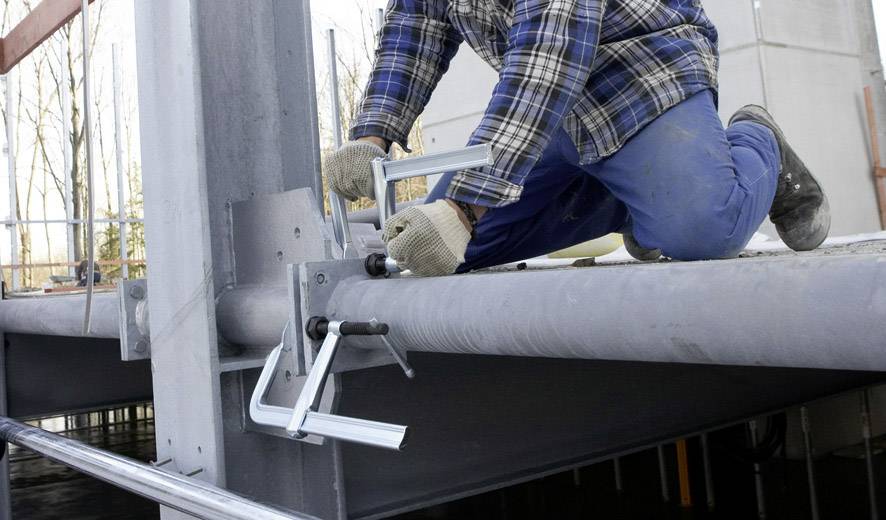 Eine Person in blauer Arbeitskleidung kniet auf einem Stahlträger und befestigt einen Metallwinkel mithilfe von Schraubzwingen.