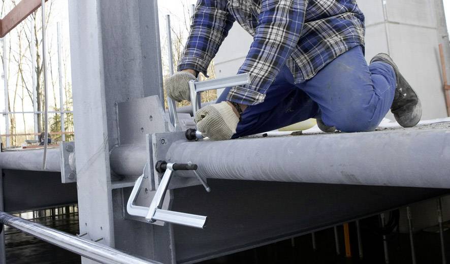 Eine Person kniet auf einem Stahlträger und befestigt Metallklemmen. Sie trägt Arbeitskleidung und Handschuhe, was auf eine Baustelle hinweist.