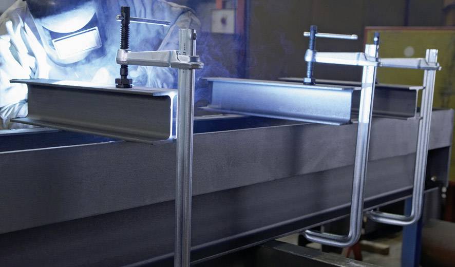 Ein Metallarbeiter schweißt zwei Stahlträger zusammen. Die Träger sind durch Klemmen fixiert. Schutzkleidung und ein heller Lichtstrahl sind sichtbar.