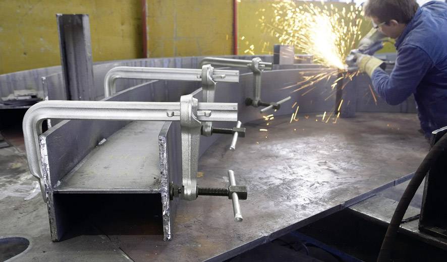 Eine Person schleift ein Metallwerkstück, wobei Funken fliegen. Im Vordergrund sind Metallklemmen an einem großen Stahlträger befestigt.
