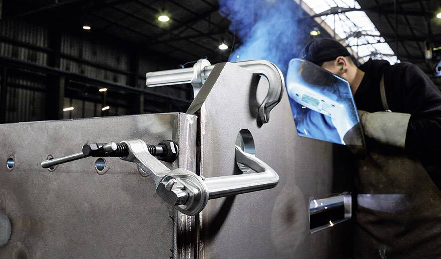 Ein Schweißer bearbeitet ein großes Metallteil in einer Fabrikhalle. Blaue Funken sprühen, und es sind Zwingen zur Stabilisierung angebracht.