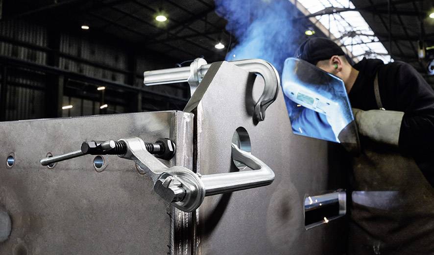 Ein Schweißer arbeitet konzentriert an einem großen Metallteil in einer Fabrikhalle. Funken fliegen, während er seine Schutzausrüstung trägt.