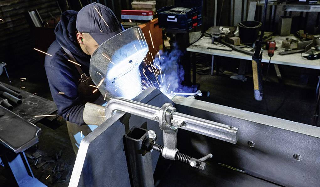 Eine Person in Schutzkleidung schweißt Metall in einer Werkstatt, Funken fliegen. Arbeitsumgebung mit Werkzeugen im Hintergrund.