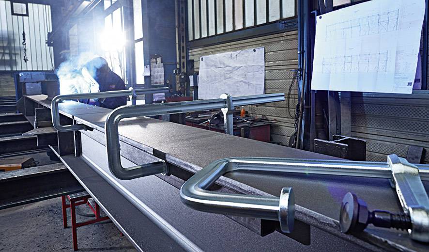 Eine Person in Schutzkleidung führt Schweißarbeiten an einem großen Metallträger in einer industriellen Werkstatt durch.