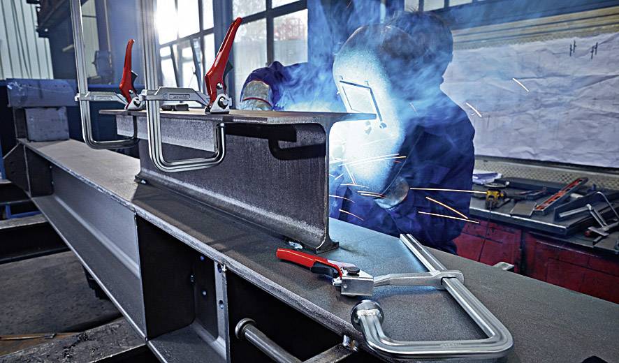 Ein Arbeiter schweißt ein Metallstück in einer Werkstatt. Funken fliegen, und rote Klemmen sichern das Werkstück auf einem Tisch.