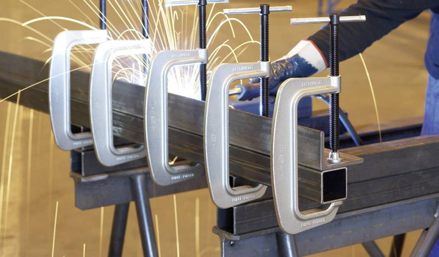 Nahaufnahme von fünf Schraubzwingen, die Metallstreben in einer Werkstatt fixieren, während Funken im Hintergrund bei Schweißarbeiten sprühen.