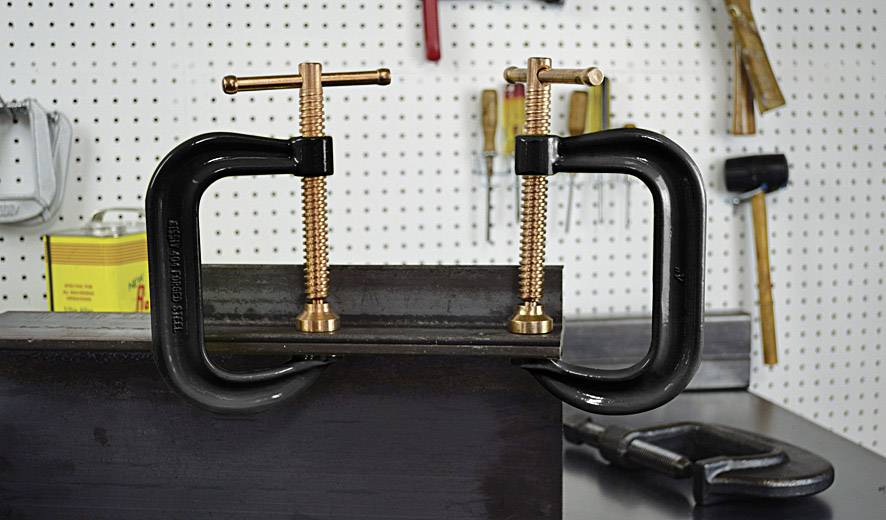 Schraubzwingen halten ein Metallstück in einer Werkstatt fest. Hintergrund zeigt Werkzeuge an der Wand.