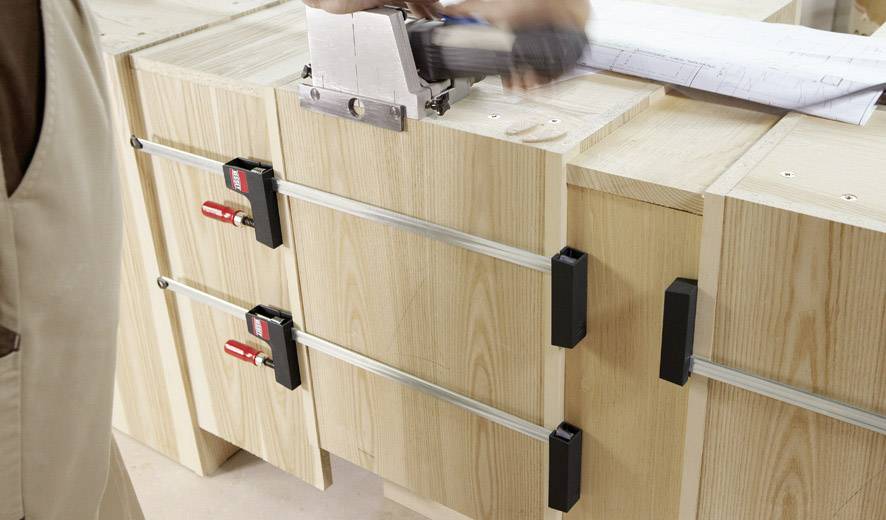 Person arbeitet an einem Holzprojekt, verwendet eine Klemme, um Holz auf einer Werkbank zu fixieren, mit Plänen im Hintergrund.