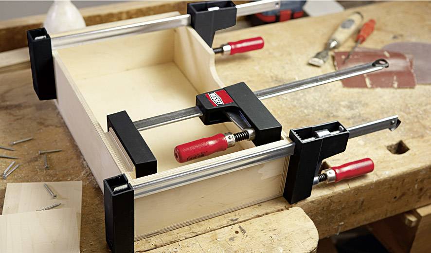 Holzwerkbank mit einer fast fertigen Schublade, die mit Schraubzwingen fixiert ist. Es sind Werkzeuge und Nägel verteilt sichtbar.