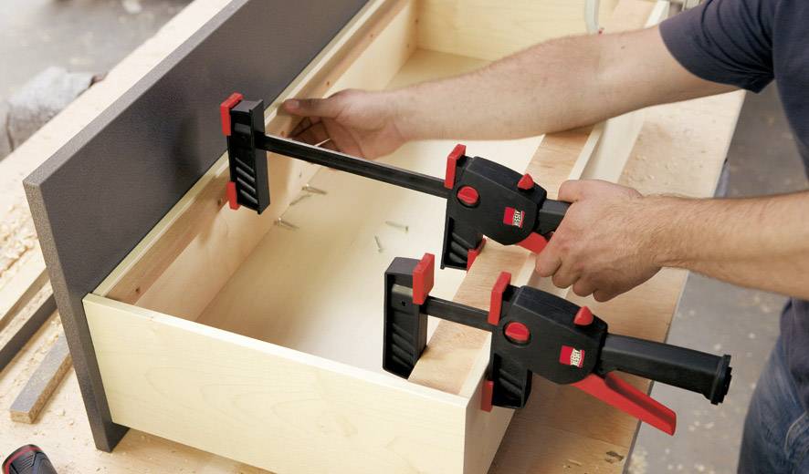 Eine Person befestigt mit Klemmen ein Holzbrett. Die Szene zeigt einen handwerklichen Arbeitsprozess in einer Werkstattumgebung.