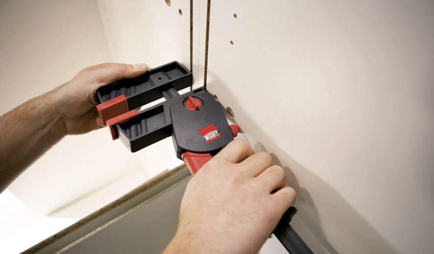 Zwei Hände verwenden eine Schraubzwinge, um Materialien zusammenzuhalten. Hintergrund aus Holz mit Bohrlöchern sichtbar. Werkzeug wird eingestellt.