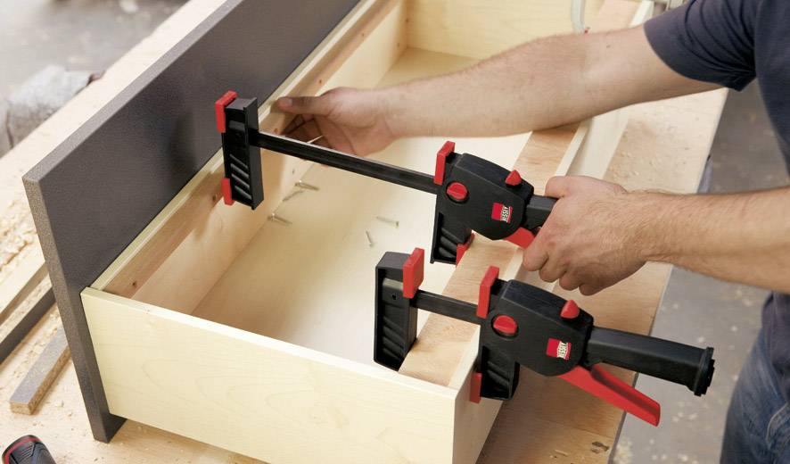 Eine Person verwendet zwei Schraubzwingen, um Holzplatten zusammenzuhalten, vermutlich im Rahmen einer Tischlerarbeit.