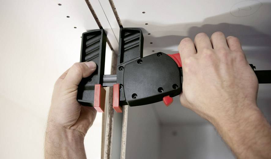 Zwei Hände halten eine Klemme, die eine Metallstange zwischen zwei Platten fixiert, um sie zusammenzudrücken und zu befestigen.