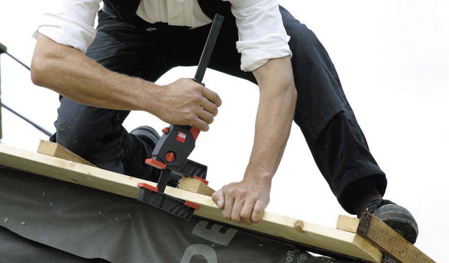 Eine Person arbeitet auf einem Dach, verwendet eine Rohrzange, um ein Holzbrett zu sichern. Sie trägt Arbeitskleidung.