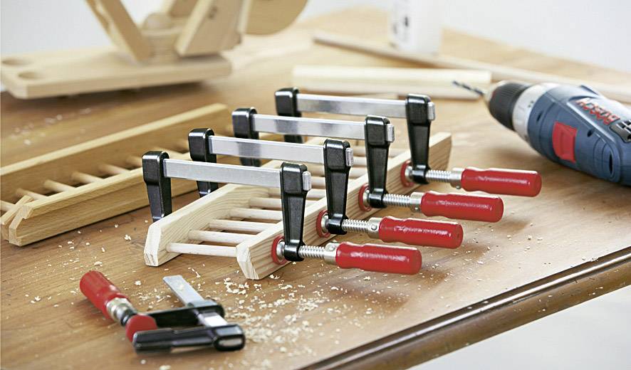An einer Holzkonstruktion auf einem Tisch werden mehrere Schraubzwingen befestigt; daneben eine Bohrmaschine und Holzspäne.