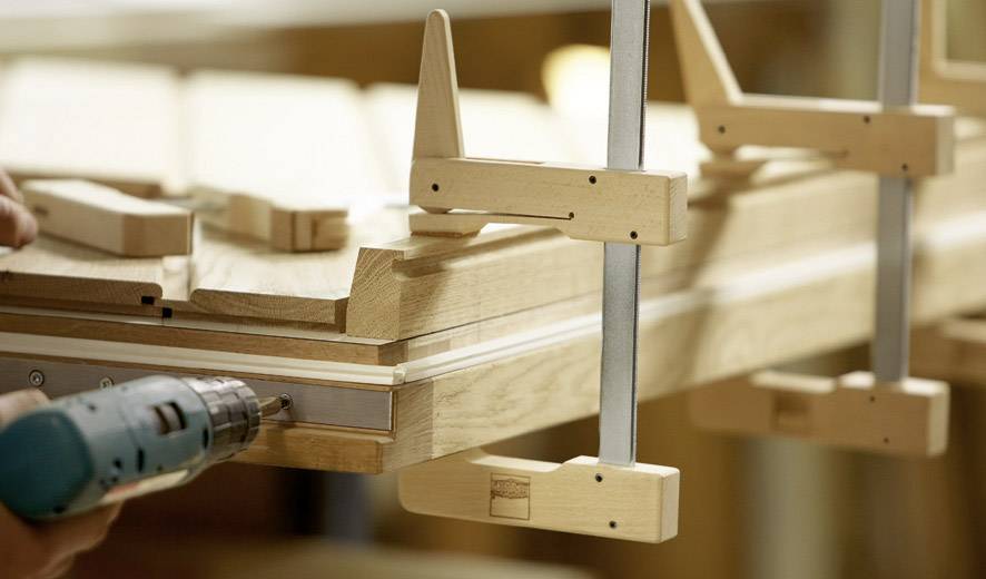 Eine Person benutzt eine Bohrmaschine, um Holzplatten zusammenzubauen, die mit Schraubzwingen fixiert sind.