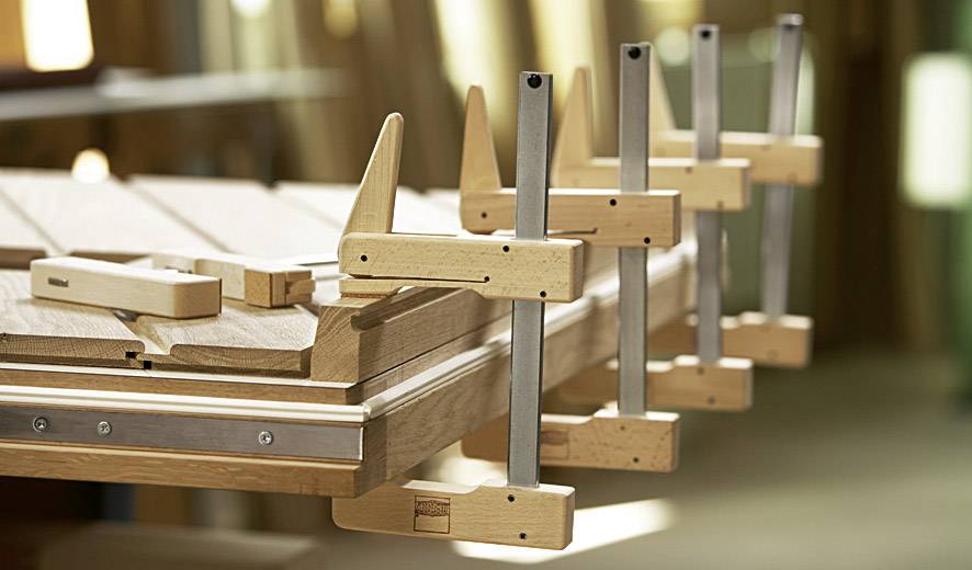 Holzlatten werden mit mehreren Schraubzwingen auf einer Werkbank fixiert. Der Raum im Hintergrund hat eine warme Holzausstattung.