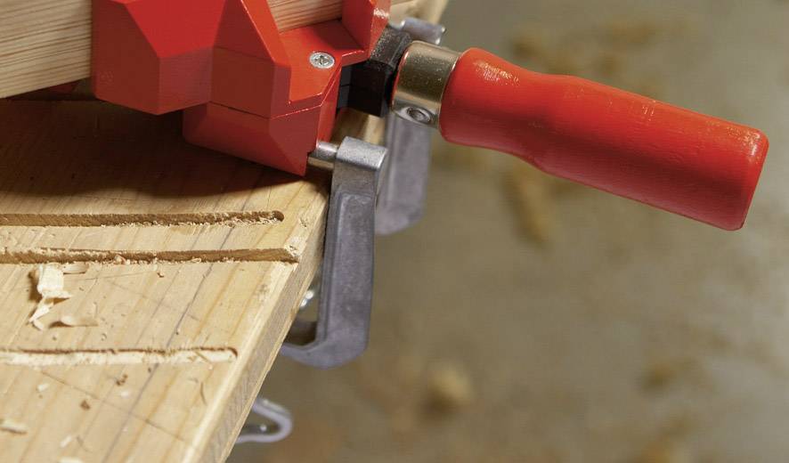 Holztischkante in einer Werkstatt. Eine rote Schraubzwinge hält Holz fest. Holzspäne liegen verteilt.