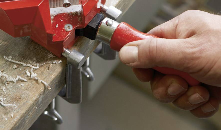 Eine Hand hält einen roten Schraubendreher, der an einem Schraubstock befestigt ist. Holzspäne liegen auf einem Holztisch.