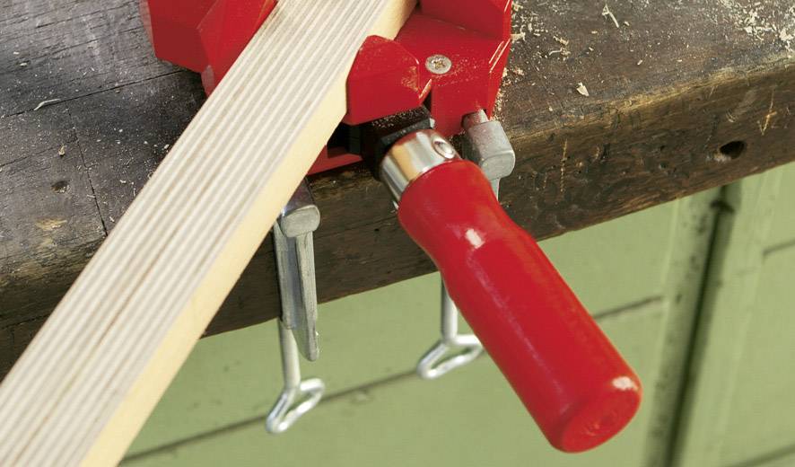 Ein Holzstück wird in einer roten Schraubzwinge auf einer Werkbank festgeklemmt. Die Zwinge hat einen roten Griff.