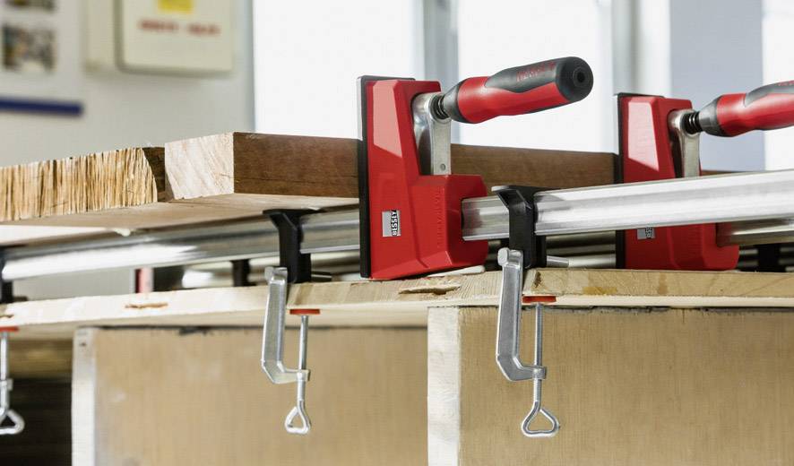 Ein Holzbrett wird mit zwei roten Schraubzwingen auf einem Tisch fixiert, möglicherweise in einer Werkstattumgebung.