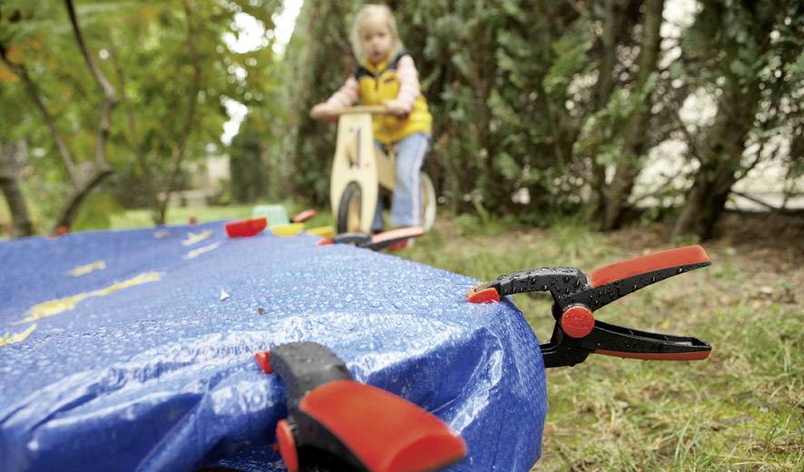 Ein Kind im Hintergrund spielt auf einem Holzspielzeug, während im Vordergrund ein blauer Stoff mit Klammern an einem Tisch befestigt ist.