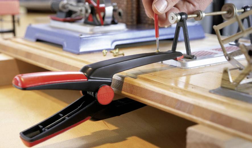 Eine schwarze und rote Klemme hält ein Holzbrett auf einem Tisch, während eine Hand ein kleines Metallteil am Brett befestigt.