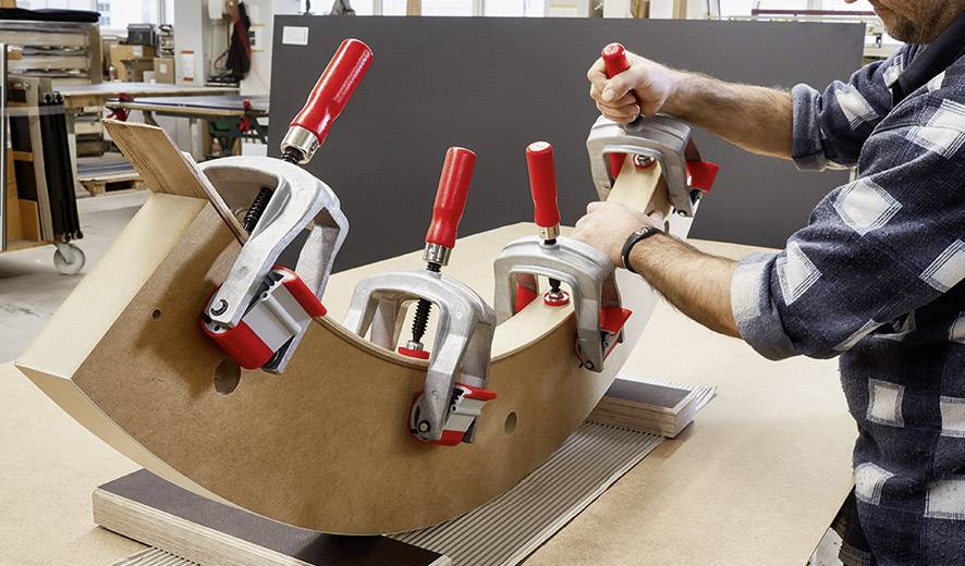 Ein Handwerker bearbeitet ein gebogenes Holzstück, das mit mehreren Klemmen auf einem Tisch befestigt ist, in einer Werkstatt.