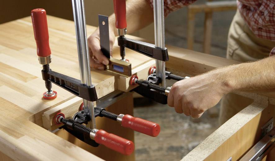 Zwei Hände, die Holz mit mehreren Schraubzwingen auf einer Werkbank fixieren. Eine Wasserwaage überprüft die Ausrichtung.
