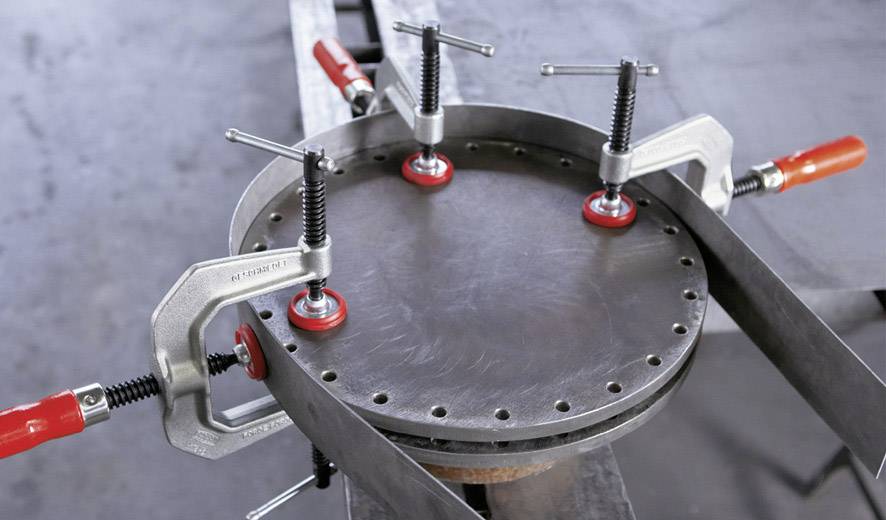 Eine metallene, runde Oberfläche wird mit vier Schraubzwingen auf einem Tisch fixiert. Die Zwingen haben rote Griffe.