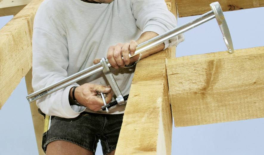 Eine Person befestigt Balken mit einem großen Metallspanner. Die Szene zeigt Holzverarbeitung oder Bauarbeiten an einem Holzkonstrukt.