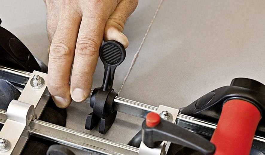 Eine Hand justiert den Hebel eines Werkzeugs, möglicherweise eines Fliesenschneiders, auf einer Metallführungsschiene.