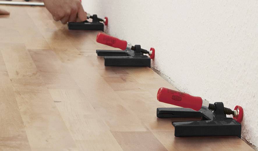 Eine Hand befestigt Dielen mit roten Plastikkeilen an einer Wand, um gleichmäßige Abstände beim Verlegen eines Holzbodens zu gewährleisten.